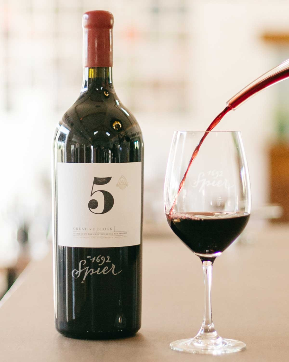 Eine Flasche Spier Creative Block 5 Rotwein steht neben einem Weinglas, das mit Rotwein gefüllt ist, auf einer Arbeitsplatte mit unscharfem Hintergrund.