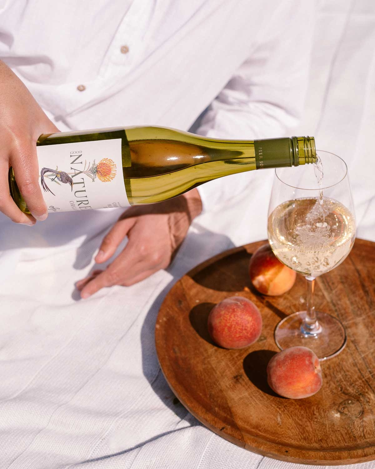 Eine Person in einem weißen Hemd gießt Weißwein aus einer Flasche in ein Glas auf einem Holztablett mit drei Pfirsichen, die alle auf einem weißen Tuch liegen.