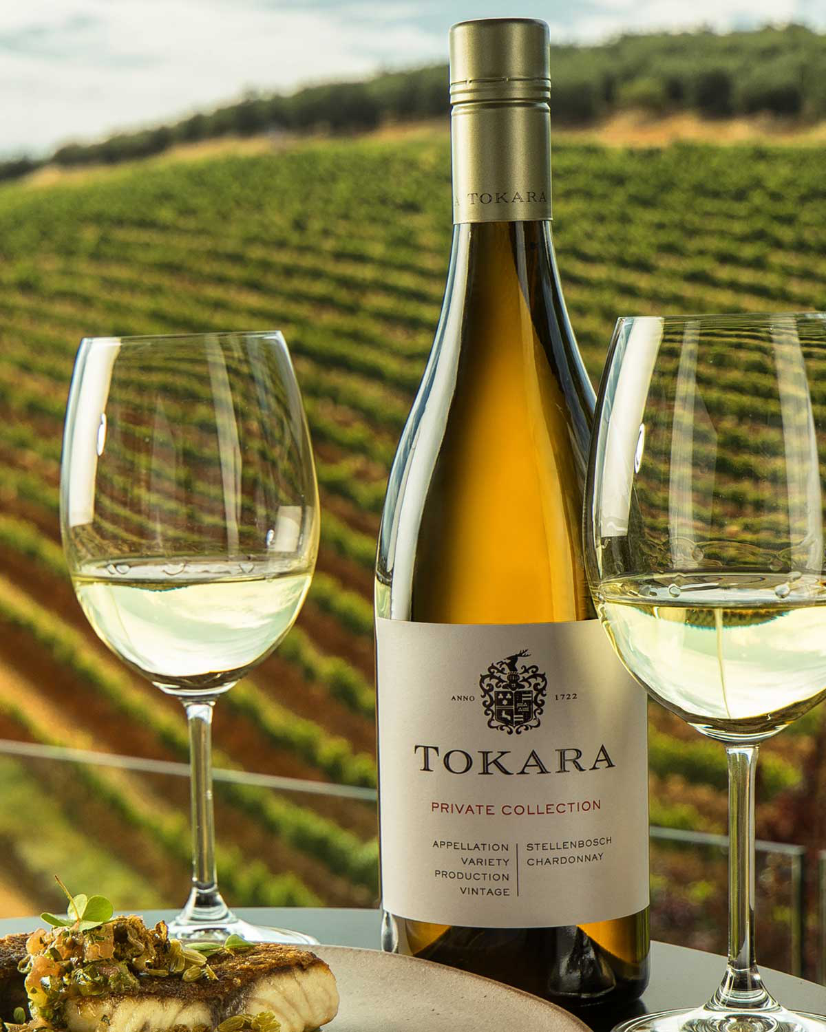 Eine Flasche Tokara Private Collection Chardonnay Wein mit einer hellen goldenen Farbe, mit einem weißen Etikett mit silbernem und goldenem Text und einem Wappen und einem passenden Tokara-gebrandeten Foliensiegel am Hals.