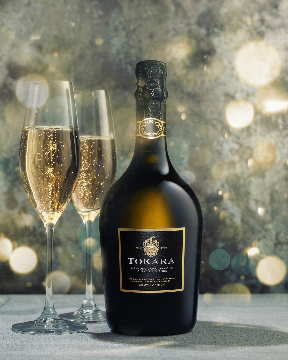 Eine Flasche Tokara-Sekt wird neben zwei gefüllten Sektflöten vor einem festlichen, verschwommenen Hintergrund mit leuchtenden Bokeh-Lichtern präsentiert.