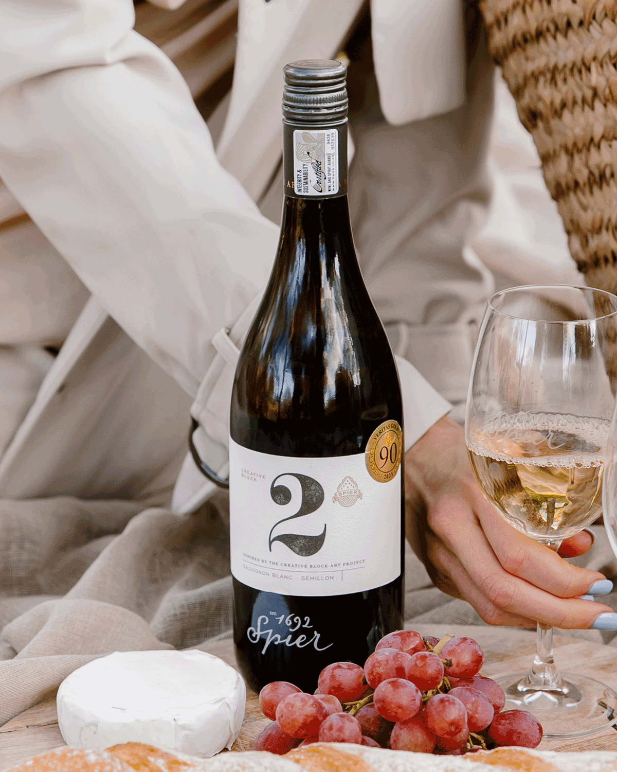 Eine Hand hält ein Glas Weißwein neben einer Flasche mit der Aufschrift "2 Spier 1692". In der Picknick-Szene sind rote Weintrauben, Brie-Käse und eine Person in einem hellen Mantel und einer gewebten Tasche zu sehen.