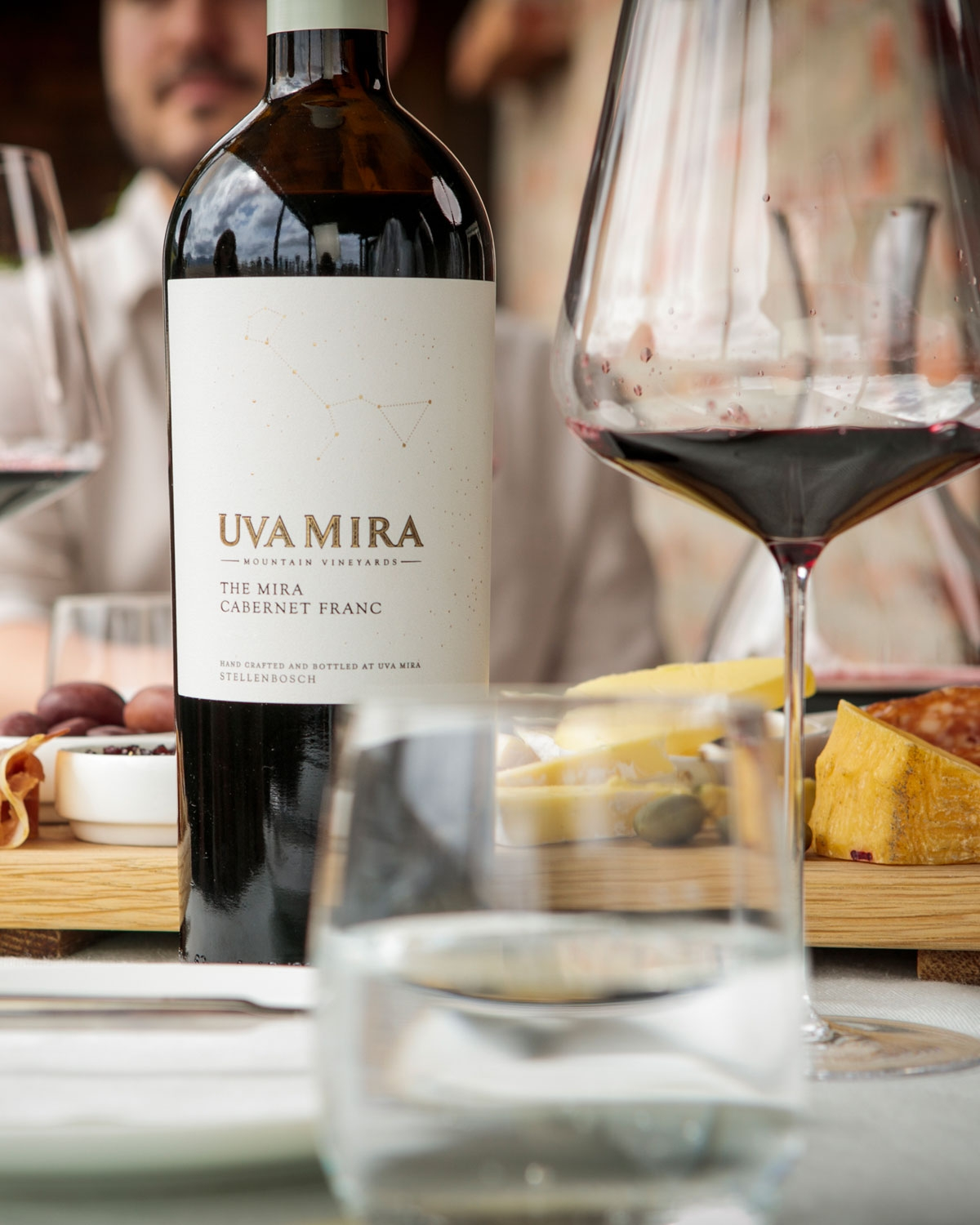 Eine Flasche des Weins Uva Mira Cabernet Franc steht auf einem Tisch mit Weingläsern, Käse, Oliven und Wurstwaren. Der unscharfe Hintergrund zeigt einen Teil einer sitzenden Person.