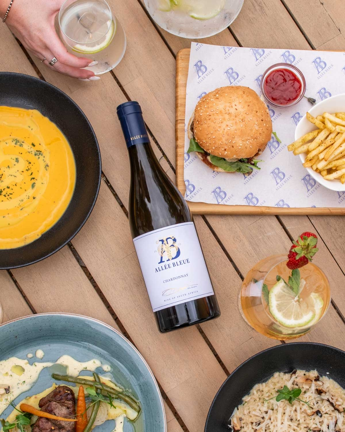 Ein gedeckter Tisch mit einem Burger und Pommes frites, einer Suppe, Nudelgerichten, einer Flasche Weißwein und zwei Getränken mit Zitrone und Garnituren. Eine Hand hält ein Glas in der Nähe der oberen linken Ecke des Bildes.