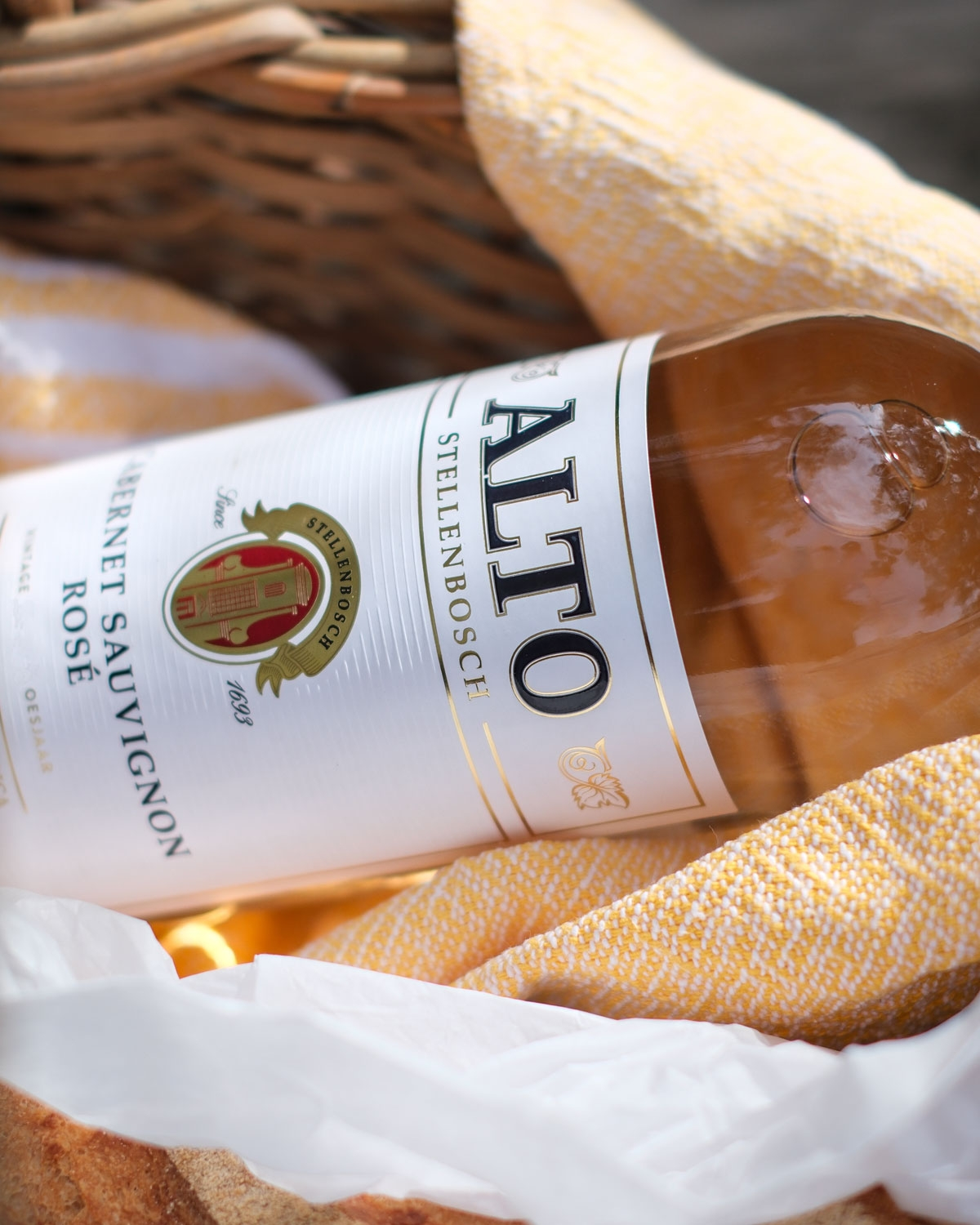 Eine Flasche Alto Stellenbosch Cabernet Sauvignon Rosé ruht in einem Weidenkorb, der mit einem gelb-weißen Tuch ausgelegt ist.