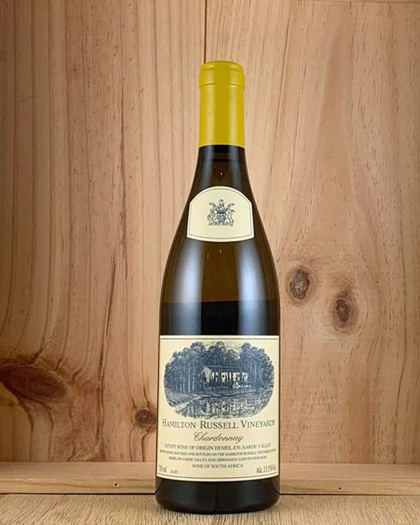 Eine Flasche Hamilton Russell Vineyards Chardonnay steht aufrecht auf einer hölzernen Oberfläche, mit einer hellgelben Kapsel und einem cremefarbenen Etikett mit Weinbergsmotiven vor einem Hintergrund aus Holzpaneelen.
