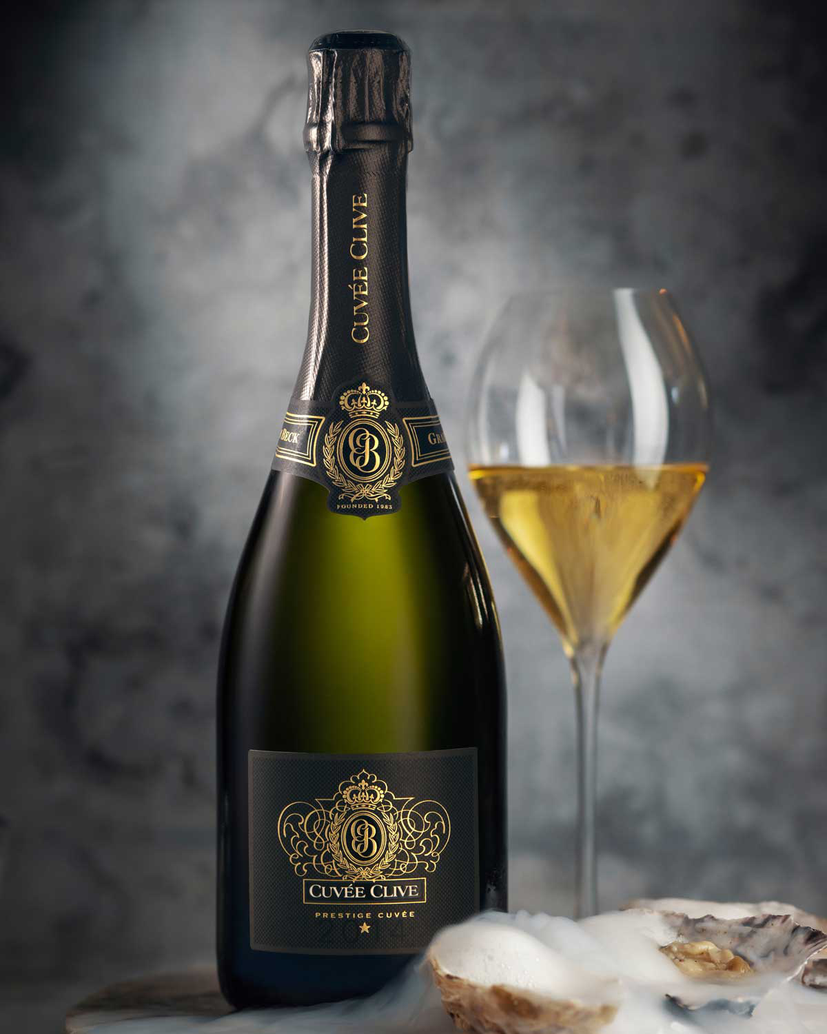 Eine Flasche Cuvée Clive Sekt steht neben einem gefüllten Weinglas, mit Austernschalen und einem Hauch von Nebel oder Schaum im Vordergrund, vor einem weichen, dunklen Hintergrund.