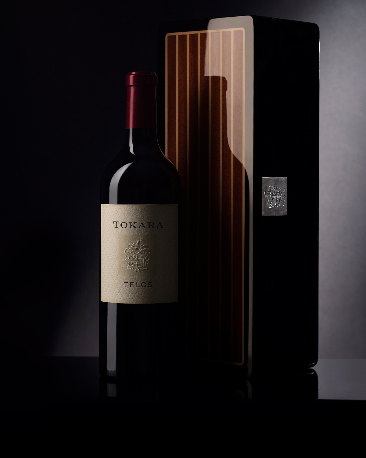 Eine Flasche Tokara Telos-Wein mit hellem Etikett steht neben einer hohen, eleganten Holzkiste mit vertikalen Intarsien, beide vor einem dunklen, sanft beleuchteten Hintergrund.