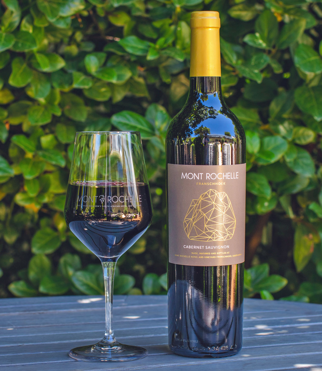 Eine Flasche Mont Rochelle Franschhoek Cabernet Sauvignon mit einem Deckel aus Goldfolie und einem dunkelbraunen Etikett mit geometrischem Goldmuster und weißem Text.