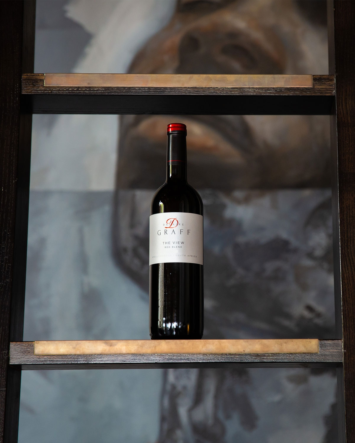 Eine Flasche Delaire Graff Rotwein steht auf einem Regal mit einem unscharfen Bild eines Gesichts im Hintergrund. Die Flasche steht zentriert und aufrecht, mit einem weißen Etikett und roter Folie am Hals.