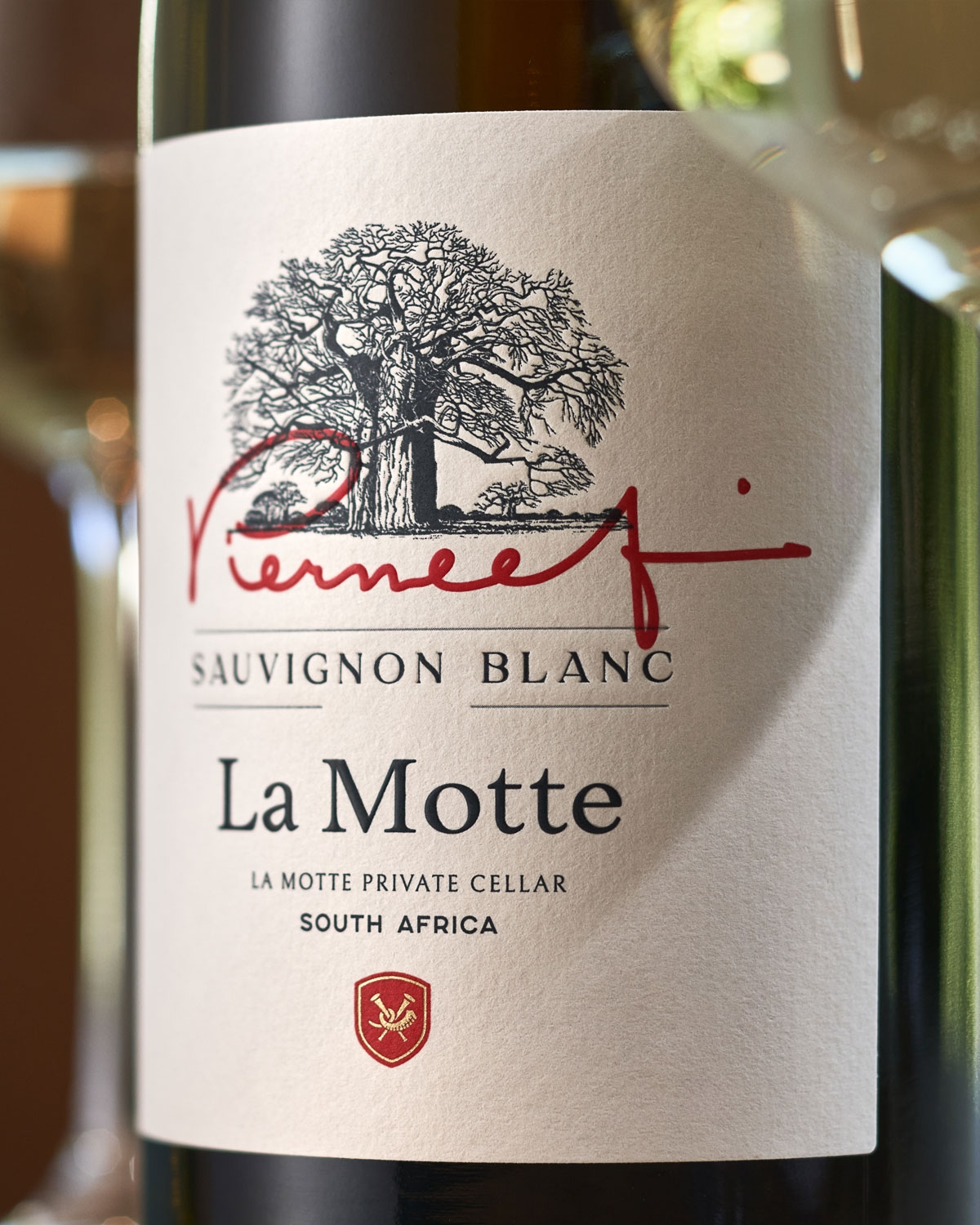 Nahaufnahme des Etiketts einer La Motte Sauvignon Blanc Weinflasche mit einer großen Baumillustration, einer roten Unterschrift und einem Text, der darauf hinweist, dass die Flasche aus einem privaten Weinkeller in Südafrika stammt. Zwei Weingläser sin