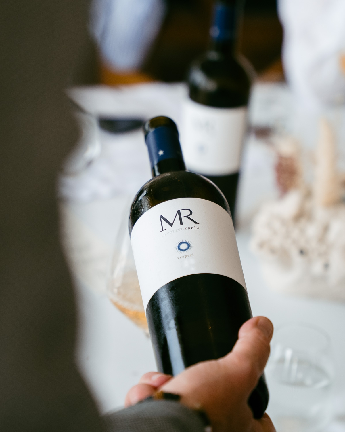 Eine Flasche Weißwein mit der Aufschrift MR und nvmveraats vesperi, mit einem minimalistischen Design mit einem weißen Etikett und einer blauen Sonnenschliff-Grafik. Die Flasche hat eine dunkelblaue Kappe mit vesperi in weißer Schrift.