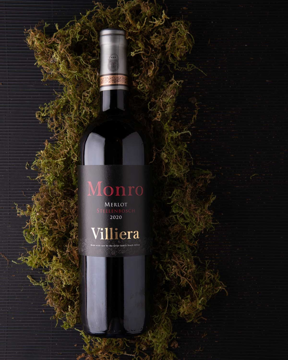 Eine Flasche Villiera Monro Merlot Wein aus Stellenbosch, Südafrika, mit einem schwarz-goldenen Folienhals, dunklem Etikett und rotem und weißem Text auf hellem Hintergrund.