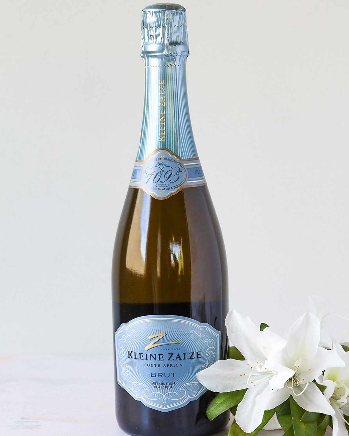 Eine Flasche Kleine Zalze Brut Schaumwein aus Südafrika steht aufrecht neben einer weißen Blume mit grünen Blättern vor einem hellen Hintergrund.