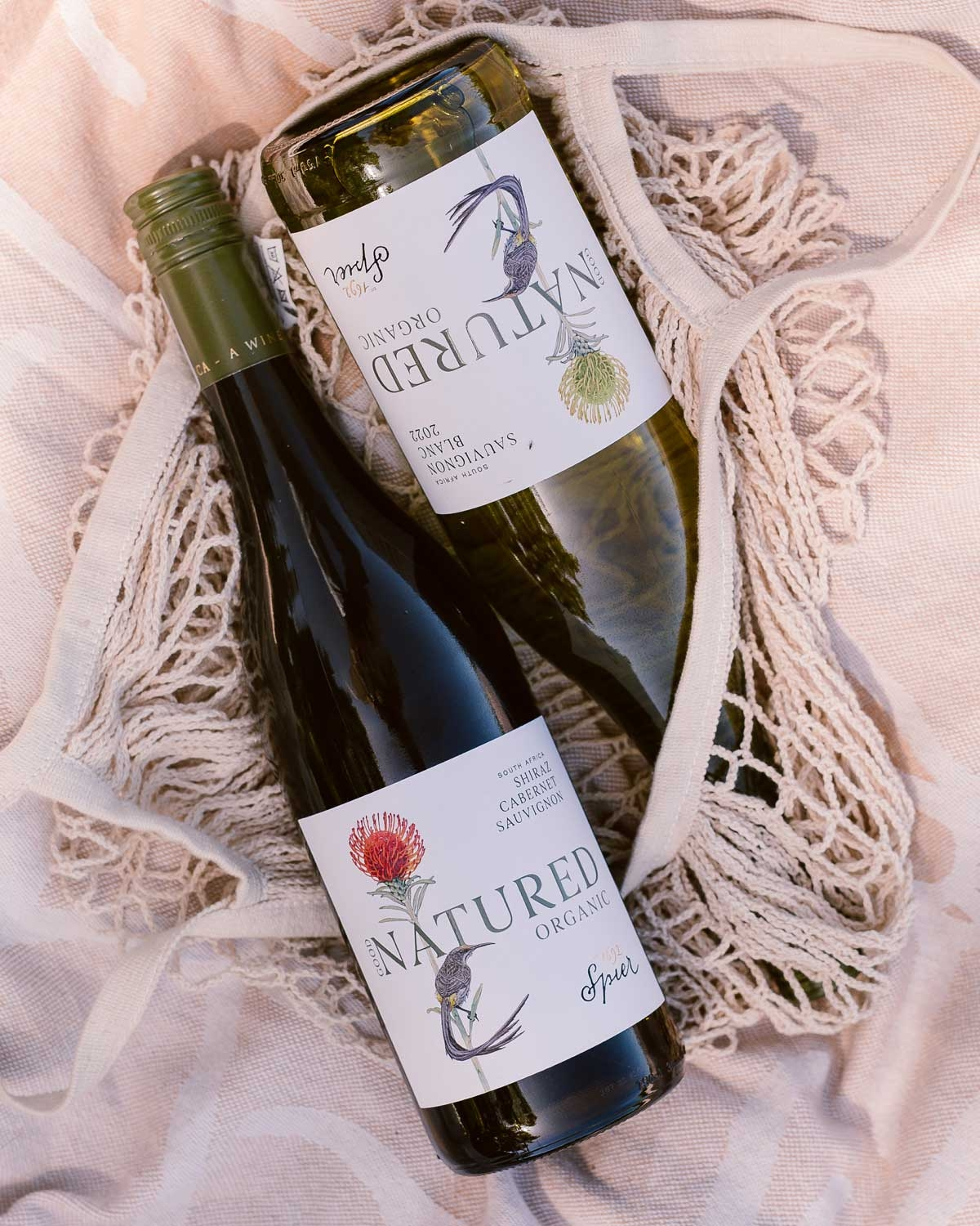 Zwei Flaschen Natured Organic Wein, eine weiße und eine rote, liegen auf einem beigefarbenen, strukturierten Tuch in einer cremefarbenen Netztasche. Auf den Etiketten beider Flaschen sind Vögel und Blumen abgebildet.