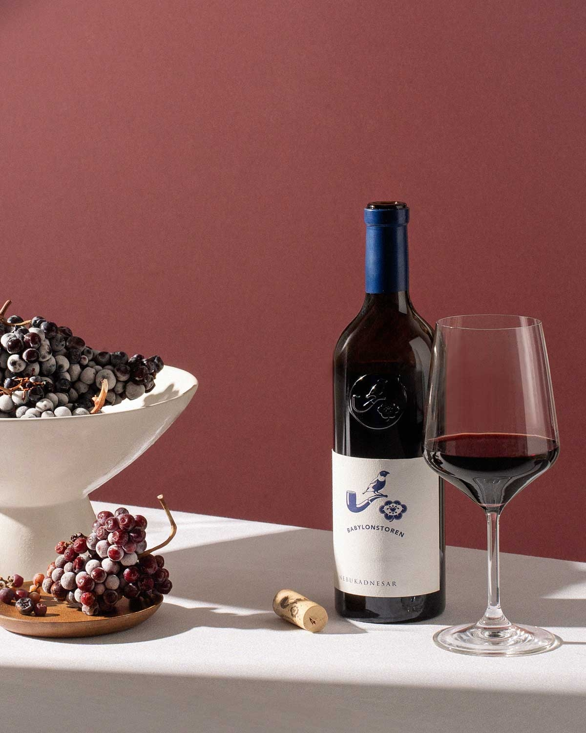 Eine Flasche Rotwein, ein gefülltes Weinglas, ein Korken und zwei Teller mit Weintrauben sind auf einer weißen Tischdecke vor einem rotbraunen Hintergrund angeordnet.