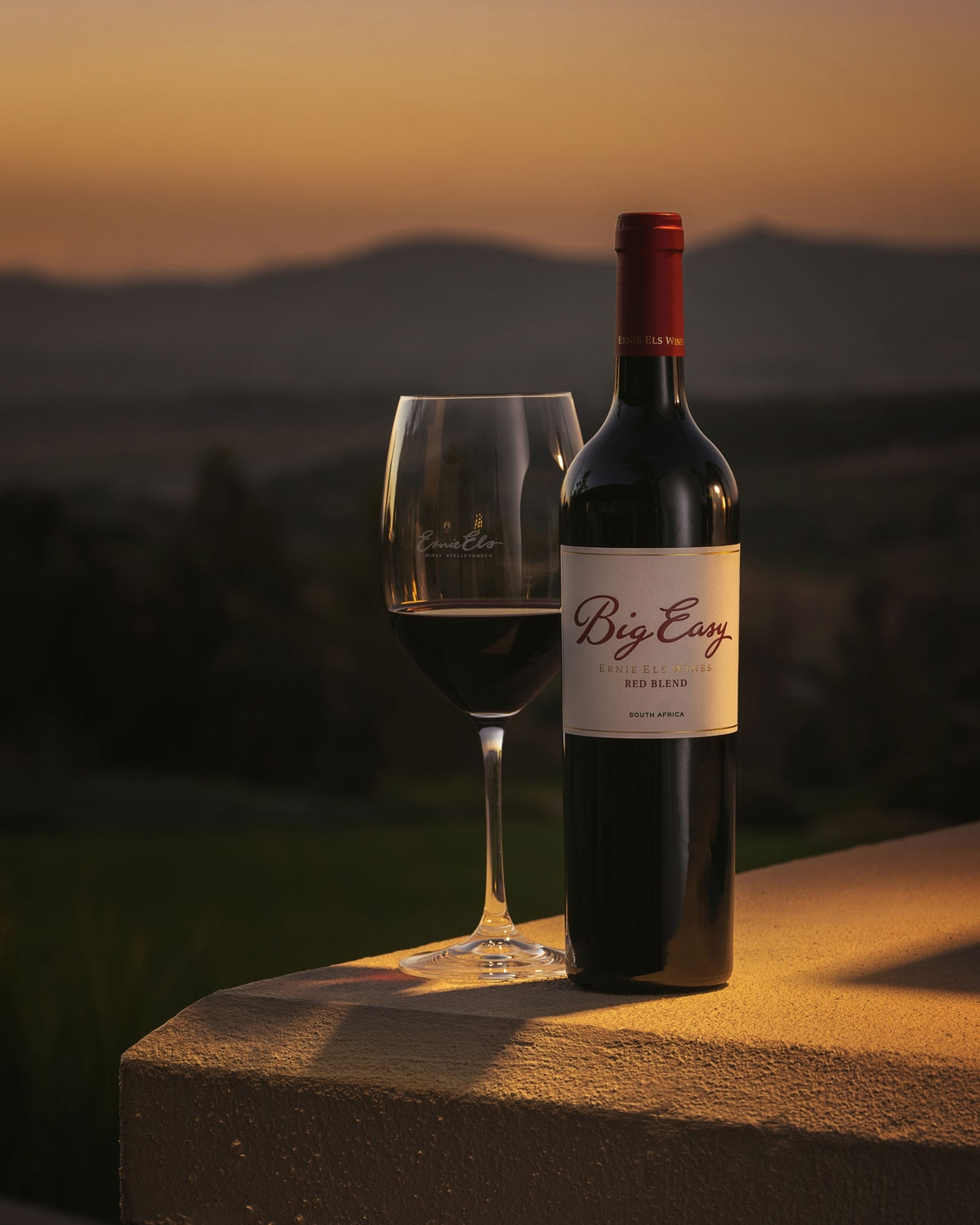 Eine Flasche Big Easy Rotwein und ein gefülltes Weinglas stehen auf einem Felsvorsprung bei Sonnenuntergang, mit sanften Hügeln und einem goldenen Himmel im Hintergrund.