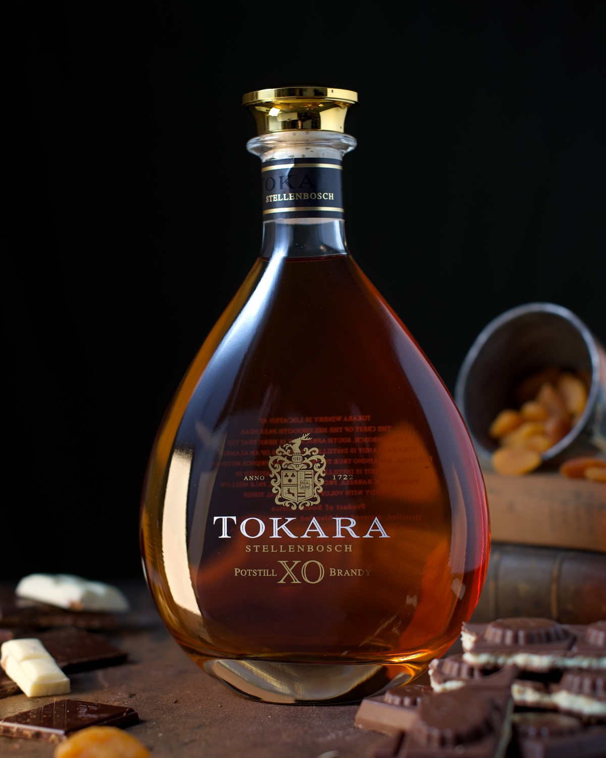 Eine Flasche Tokara XO Potstill Brandy steht auf einer dunklen Fläche, umgeben von Schokoladenstückchen und getrockneten Aprikosen, die aus einem Metallbecher quellen, alles vor einem dunklen Hintergrund.