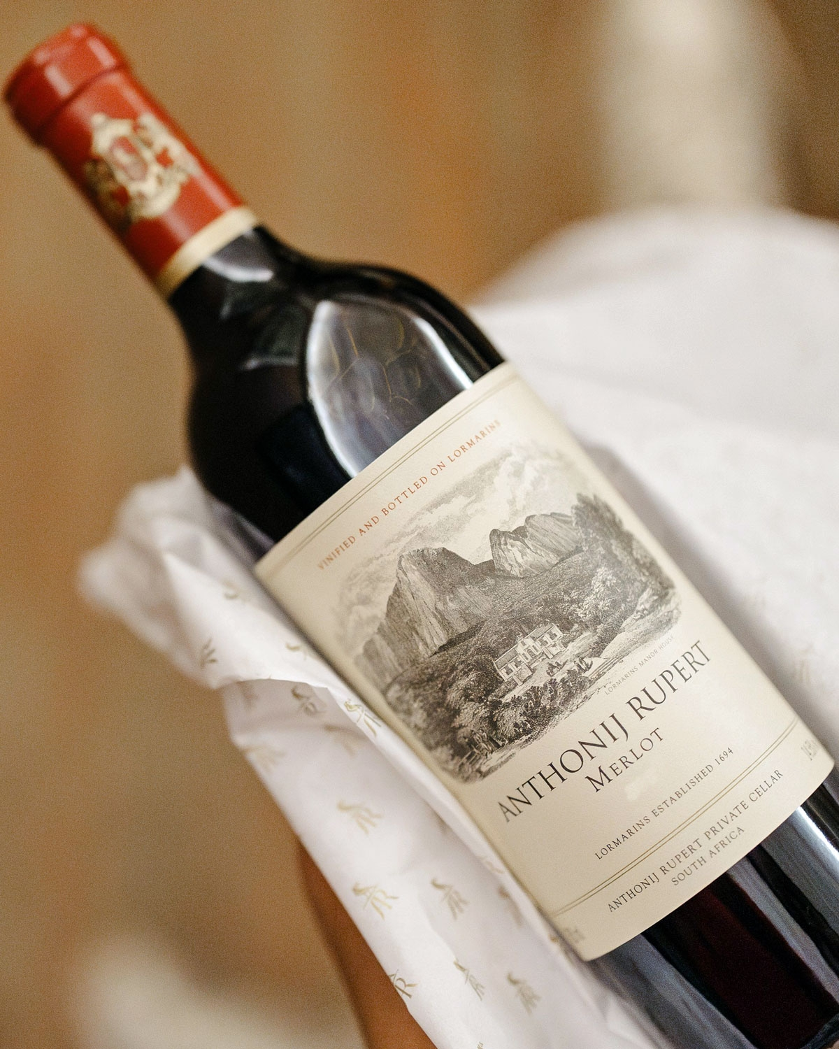 Nahaufnahme einer Flasche Anthonij Rupert Merlot-Wein mit einem detaillierten Etikett, auf dem Berge und ein Weinberg abgebildet sind und das auf weißem Seidenpapier liegt.