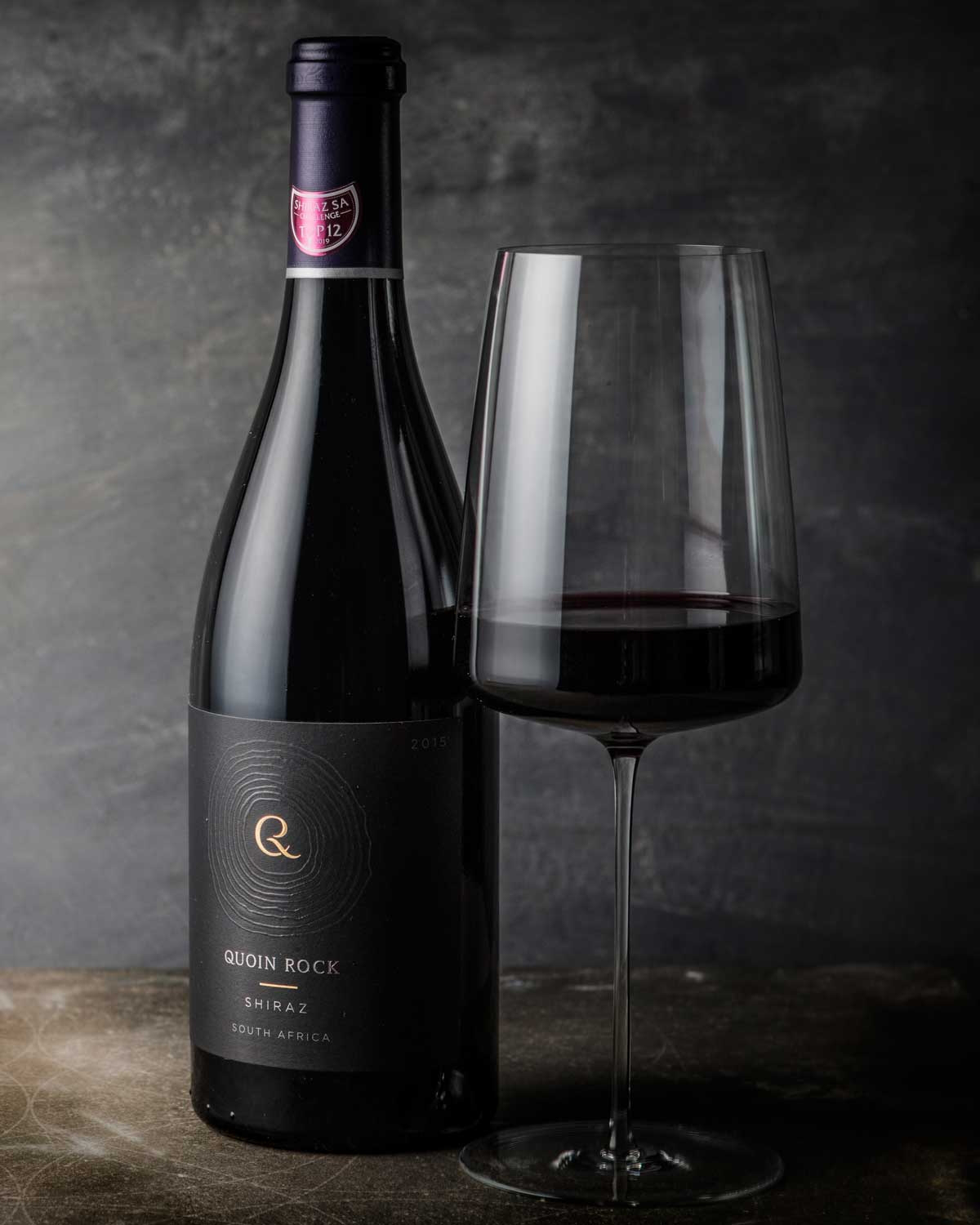 Eine dunkle Glasflasche mit dem Wein Quoin Rock Shiraz aus Südafrika, mit einem schwarzen Etikett mit goldener und weißer Schrift und einer violetten Folie auf dem Deckel.