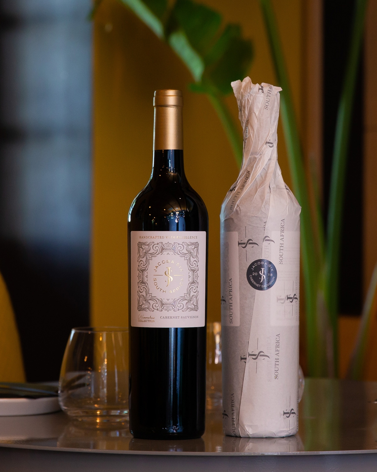 Eine Flasche Rotwein mit goldenem und weißem Etikett steht neben einer zweiten, in Markenpapier eingewickelten Flasche auf einem nachdenklichen Tisch mit leeren Weingläsern und einer grünen Pflanze im Hintergrund.