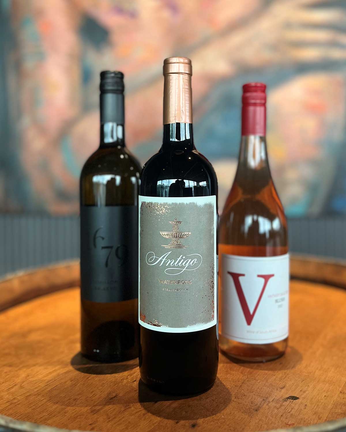 Drei Weinflaschen - eine rote, eine roséfarbene und eine dunkle - stehen auf einem Holzfass mit einem verschwommenen, bunten Gemälde im Hintergrund. Auf dem vorderen Etikett steht "Antigo".