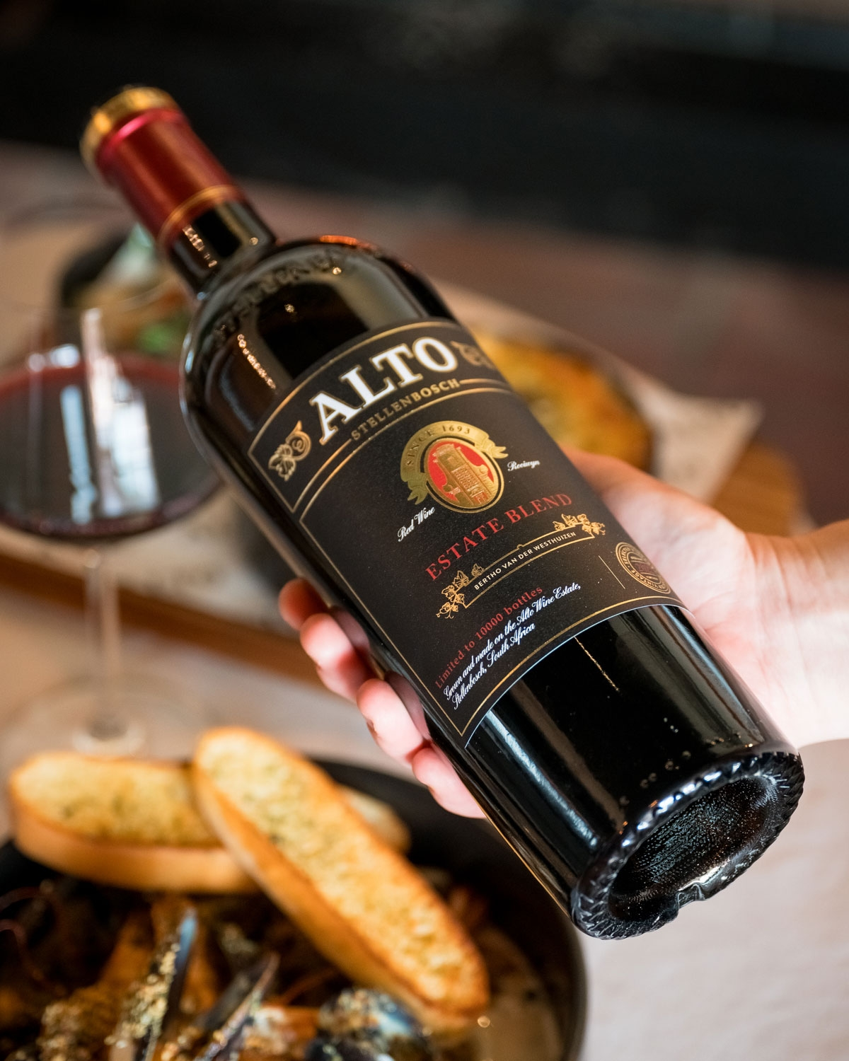 Eine Hand hält eine Flasche Alto Estate Blend Rotwein über einem gedeckten Tisch mit einem Glas Rotwein, Knoblauchbrotscheiben und einem Teller mit Speisen im Hintergrund.