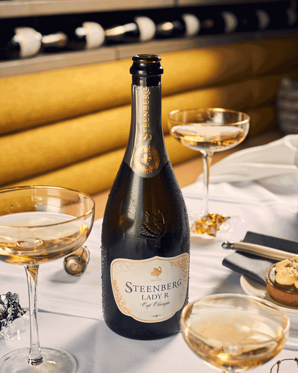 Eine Flasche Steenberg Lady R Sekt auf einem weißen Tischtuch, umgeben von drei gefüllten Champagner-Coupe-Gläsern, Pralinen und Häppchen, mit einem Weinregal und gepolsterten Sitzgelegenheiten im Hintergrund.