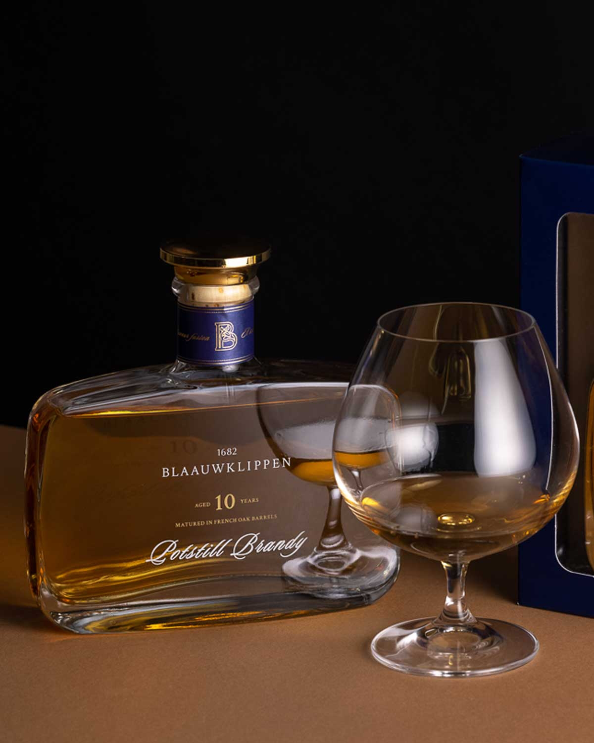 Eine Flasche Blaauwklippen 10 Jahre alter Brandy steht neben einem gefüllten Schnapsglas auf einer braunen Fläche, rechts ist ein Teil einer blauen Schachtel zu sehen.