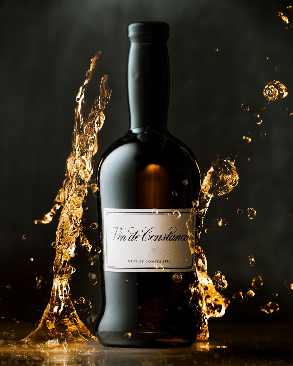 Eine Flasche Vin de Constance steht aufrecht und ist von einer goldenen Flüssigkeit umgeben, die vor einem dunklen Hintergrund dramatisch spritzt. Das Etikett auf der Flasche ist deutlich sichtbar.