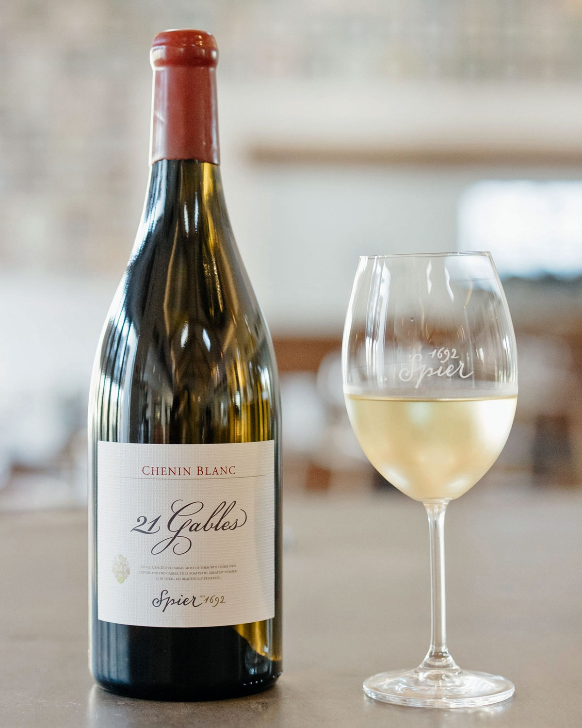Eine Flasche Spier 21 Gables Chenin Blanc steht neben einem gefüllten Weinglas auf einem Tisch, mit einem unscharfen Hintergrund einer Innenraumumgebung.