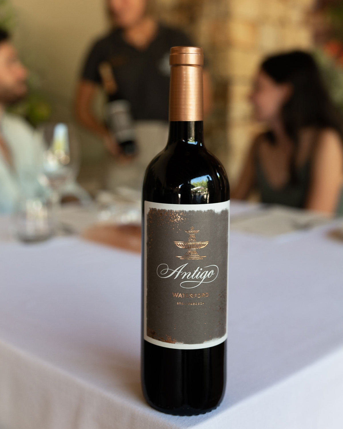 Eine Flasche Antigo-Rotwein steht auf einem weißen Tischtuch, zwei Personen sitzen und unterhalten sich im unscharfen Hintergrund, eine weitere Person steht und hält eine Weinflasche.