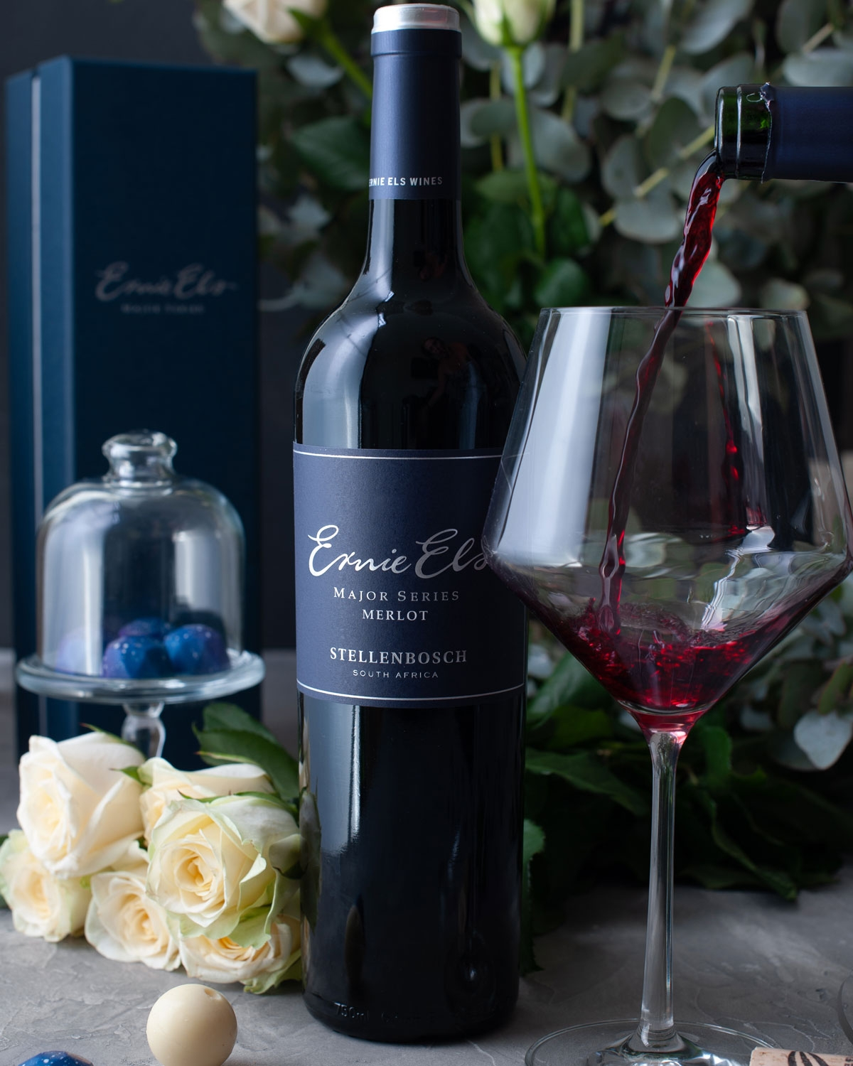Eine Flasche Ernie Els Merlot Wein steht neben einem großen Glas, das mit Rotwein gefüllt ist. Weiße Rosen, eine Schachtel, Pralinen und Grünzeug schmücken den eleganten Rahmen.