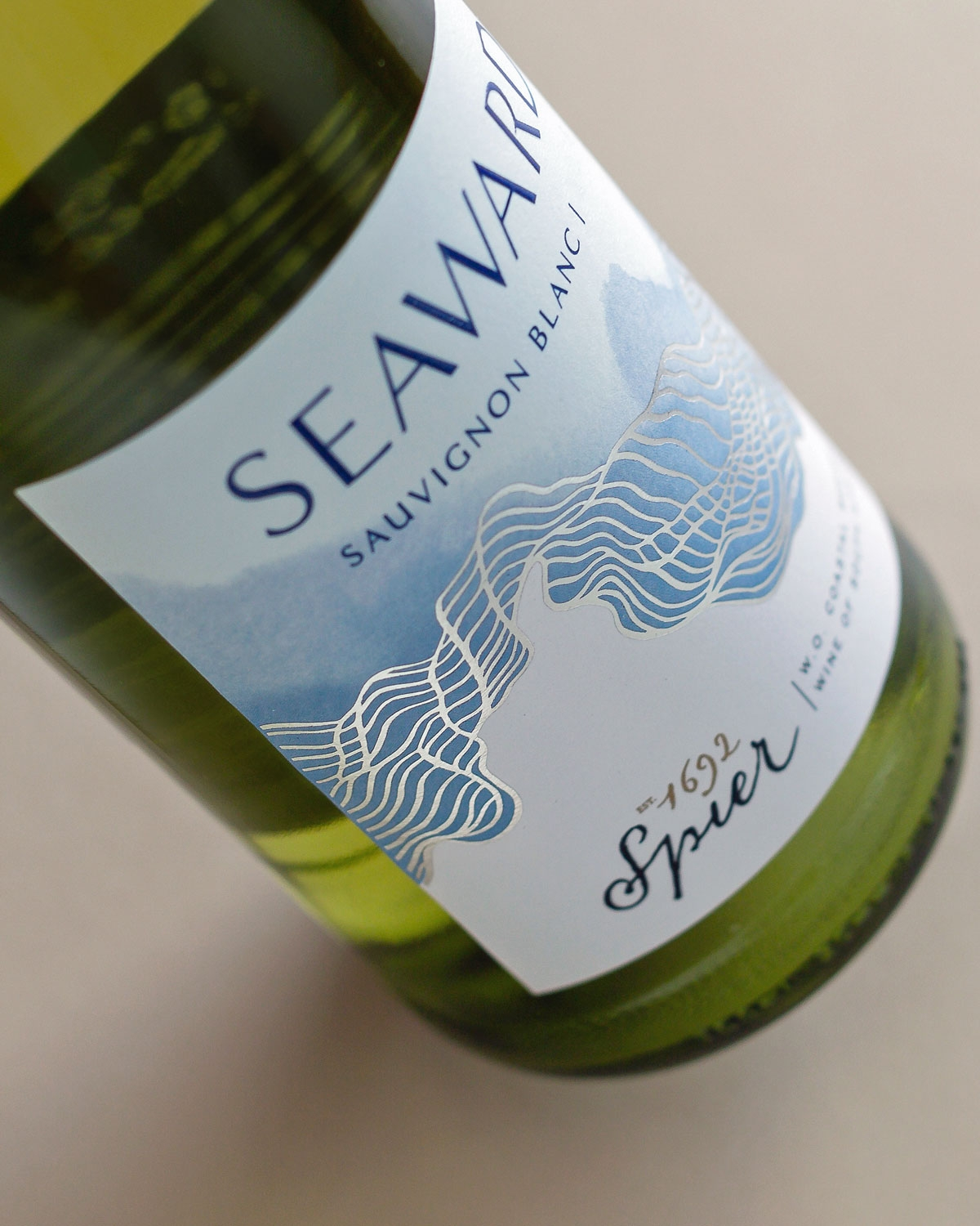 Eine Nahaufnahme einer Weinflasche mit der Aufschrift Seaward Sauvignon Blanc 1692 Spier und einem blau-weißen Wellenmuster auf dem Etikett. Die Flasche ist vor einem neutralen Hintergrund geneigt.