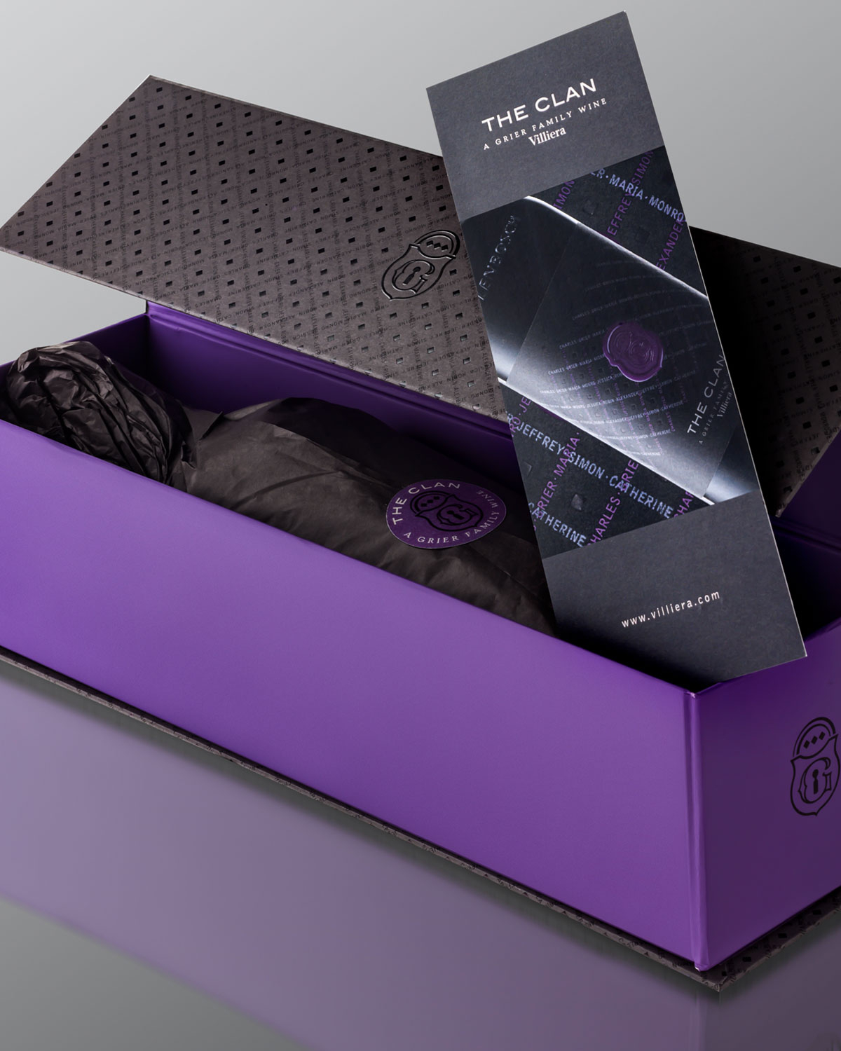 Eine dunkle Weinflasche mit der Aufschrift The Clan in einer eleganten schwarz-violetten Geschenkbox mit geometrischem Muster und geprägtem Logo, die teilweise geöffnet ist, um die Flasche zu enthüllen.