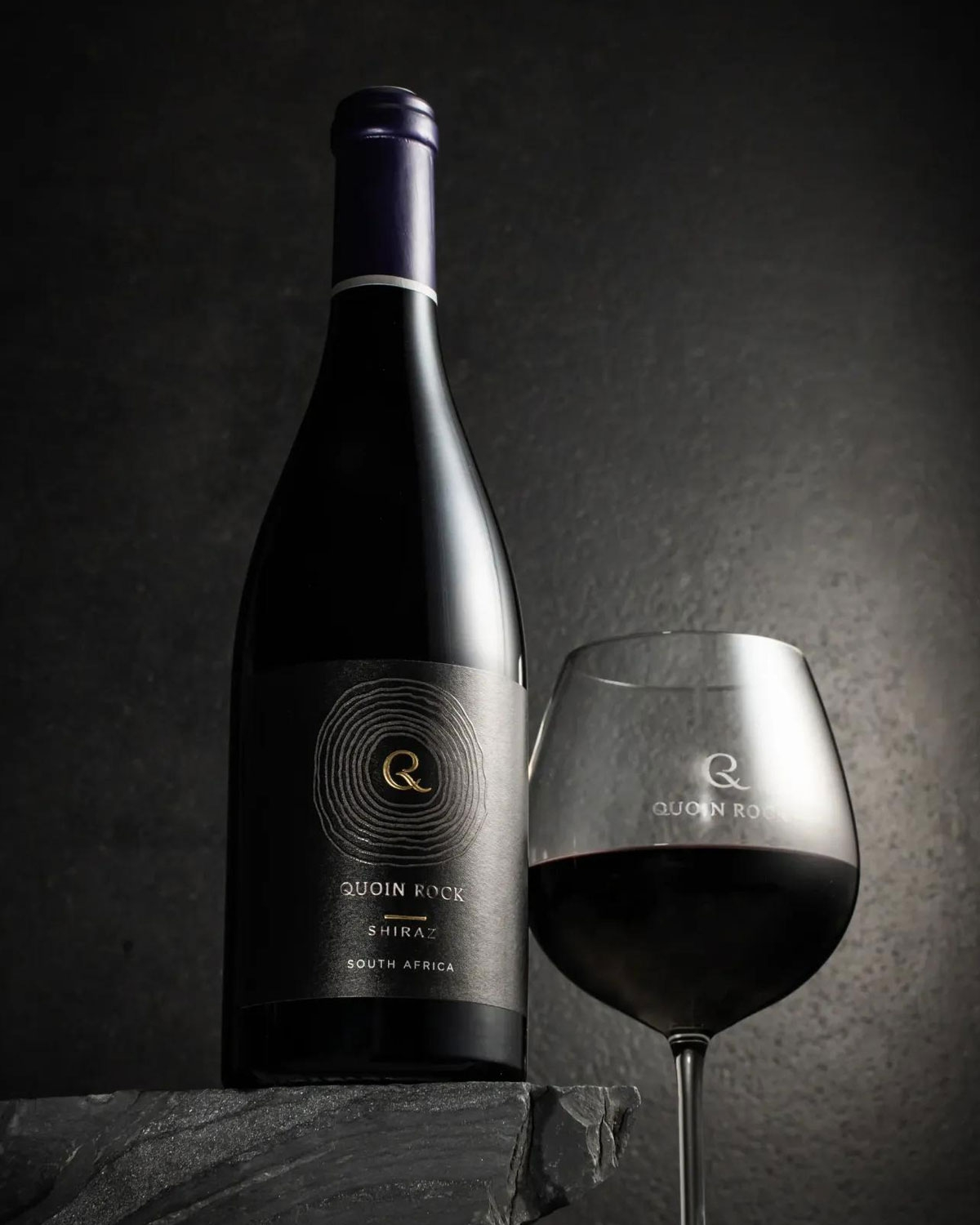 Eine Flasche Quoin Rock Shiraz Wein aus Südafrika steht neben einem gefüllten Weinglas, beide vor einem dunklen Hintergrund, auf dem das schlichte schwarz-goldene Etikett durch dezente Beleuchtung hervorgehoben wird.