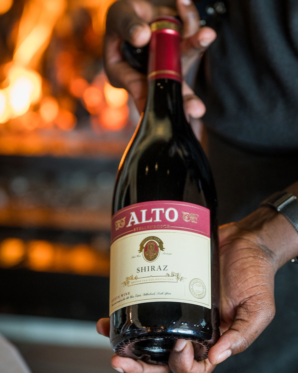 Eine Person hält eine Flasche Alto Shiraz Rotwein vor einem warmen, verschwommenen Kamin.