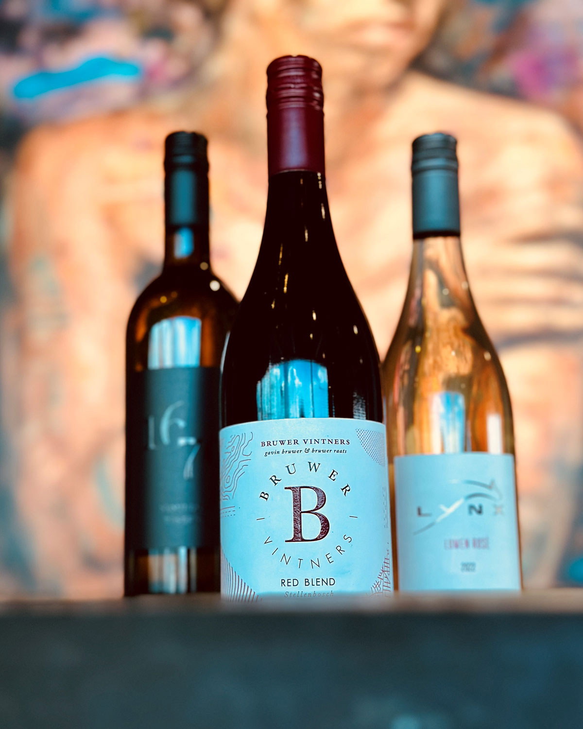 Drei Weinflaschen sind vor einem unscharfen, bunten Porträt ausgestellt. Die mittlere Flasche ist ein Bruwer Vintners Red Blend, flankiert von zwei weiteren Flaschen mit teilweise verdeckten Etiketten.
