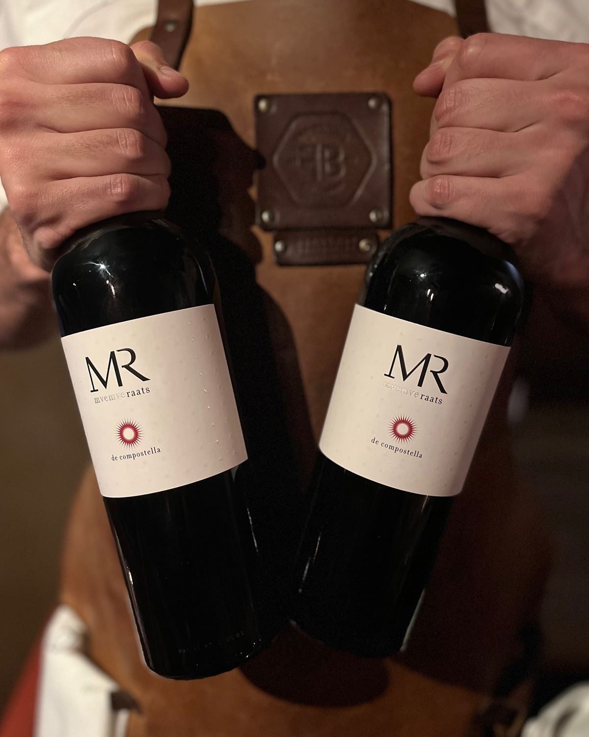 Eine Flasche Rotwein mit rotem Verschluss. Auf dem weißen Etikett steht "MR" und "De Compostella", darunter ein roter Sonnenschliff. Der Hintergrund ist einfarbig und hell gefärbt.