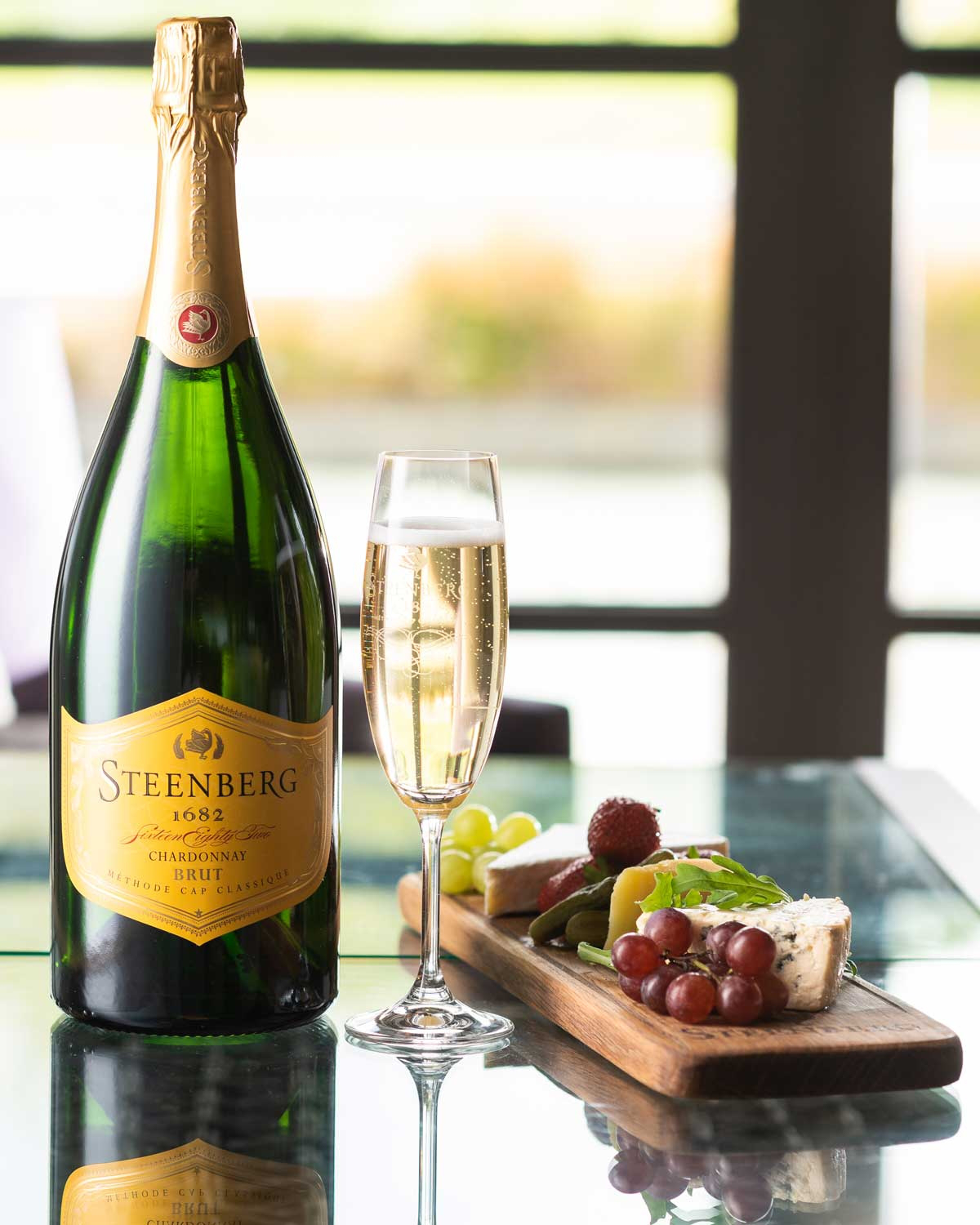 Auf einem spiegelnden Glastisch stehen eine Flasche Steenberg Chardonnay Brut, eine gefüllte Sektflöte und eine Holzplatte mit verschiedenen Käsesorten, Fleisch, Weintrauben und Grünzeug.