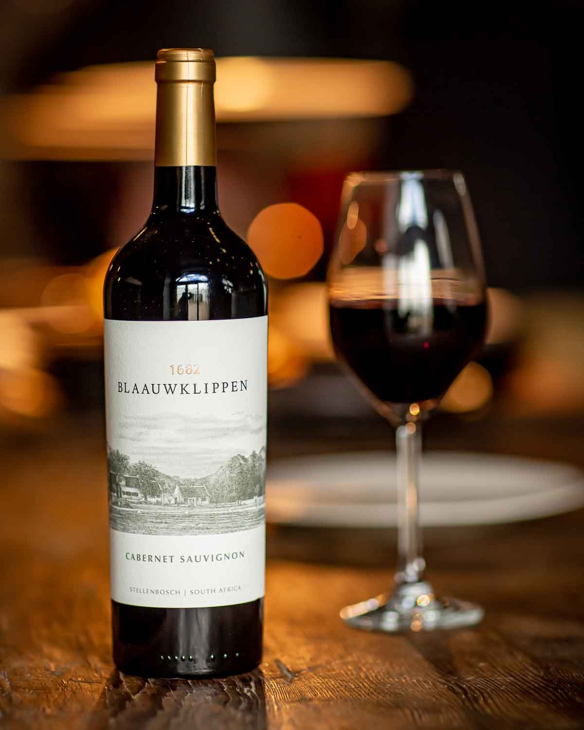Eine Flasche Blaauwklippen Cabernet Sauvignon Wein steht neben einem gefüllten Weinglas auf einem Tisch, mit einem warmen, weich verschwommenen Hintergrund.