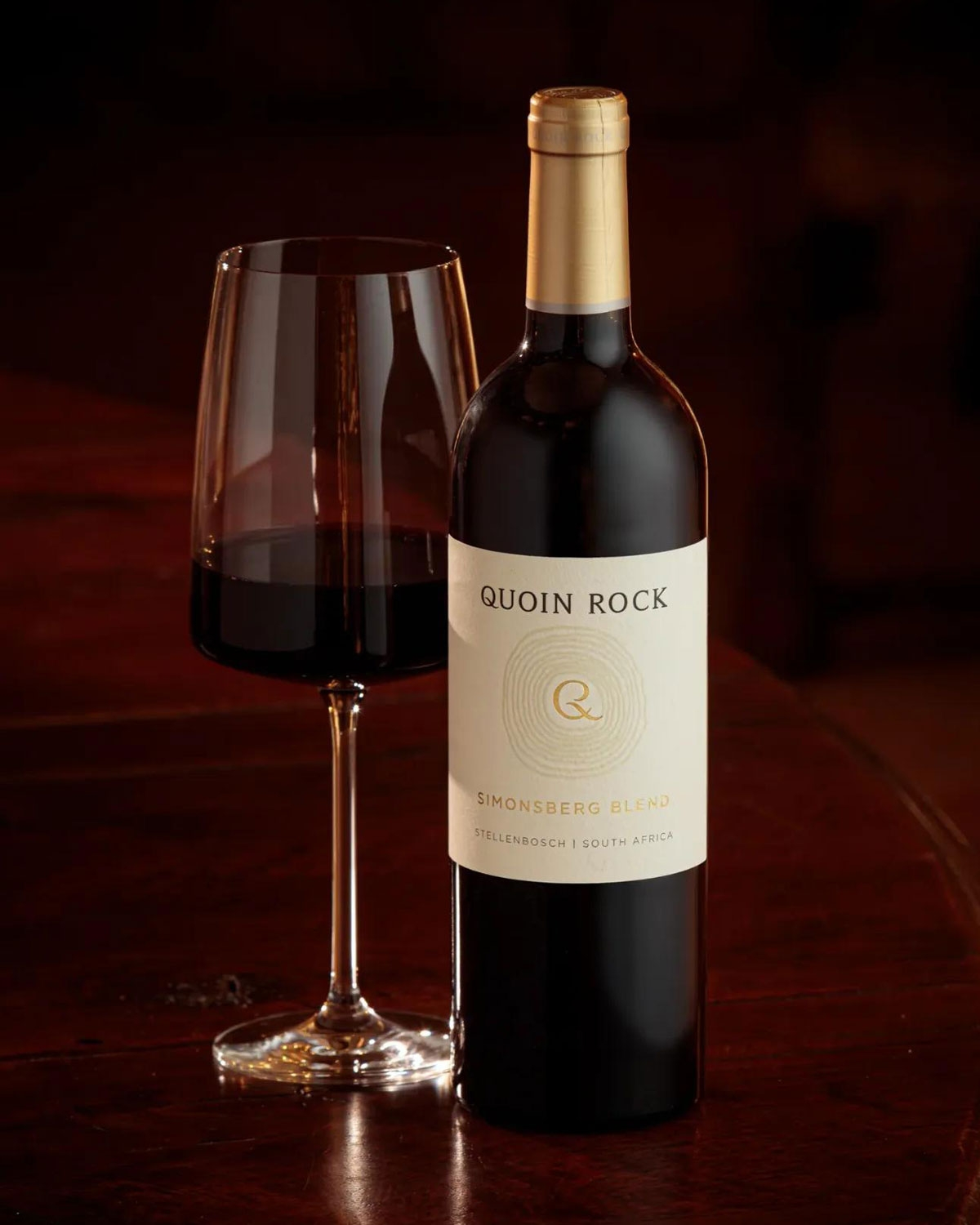Eine Flasche Quoin Rock Simonsberg Blend Rotwein aus Stellenbosch, Südafrika, mit einem cremefarbenen Etikett und goldenen Akzenten.