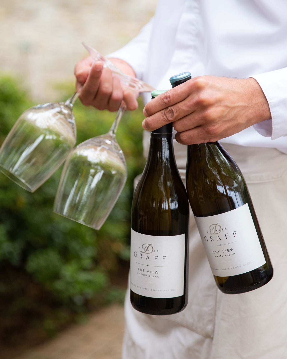 Eine Person in einem weißen Hemd hält zwei Weingläser in einer Hand und zwei Flaschen Delaire Graff Wein in der anderen, im Freien mit Grün im Hintergrund.