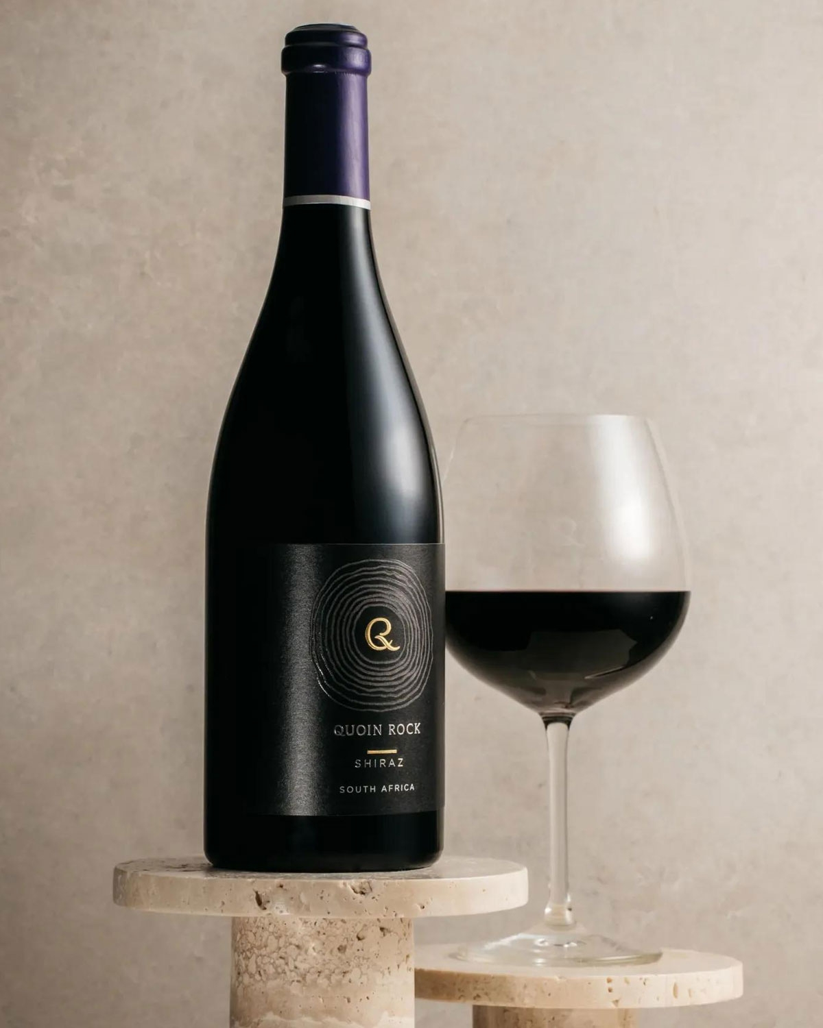 Eine Flasche Quoin Rock Shiraz Rotwein steht neben einem gefüllten Weinglas auf einer hellen Steinfläche vor einem neutralen beigen Hintergrund.