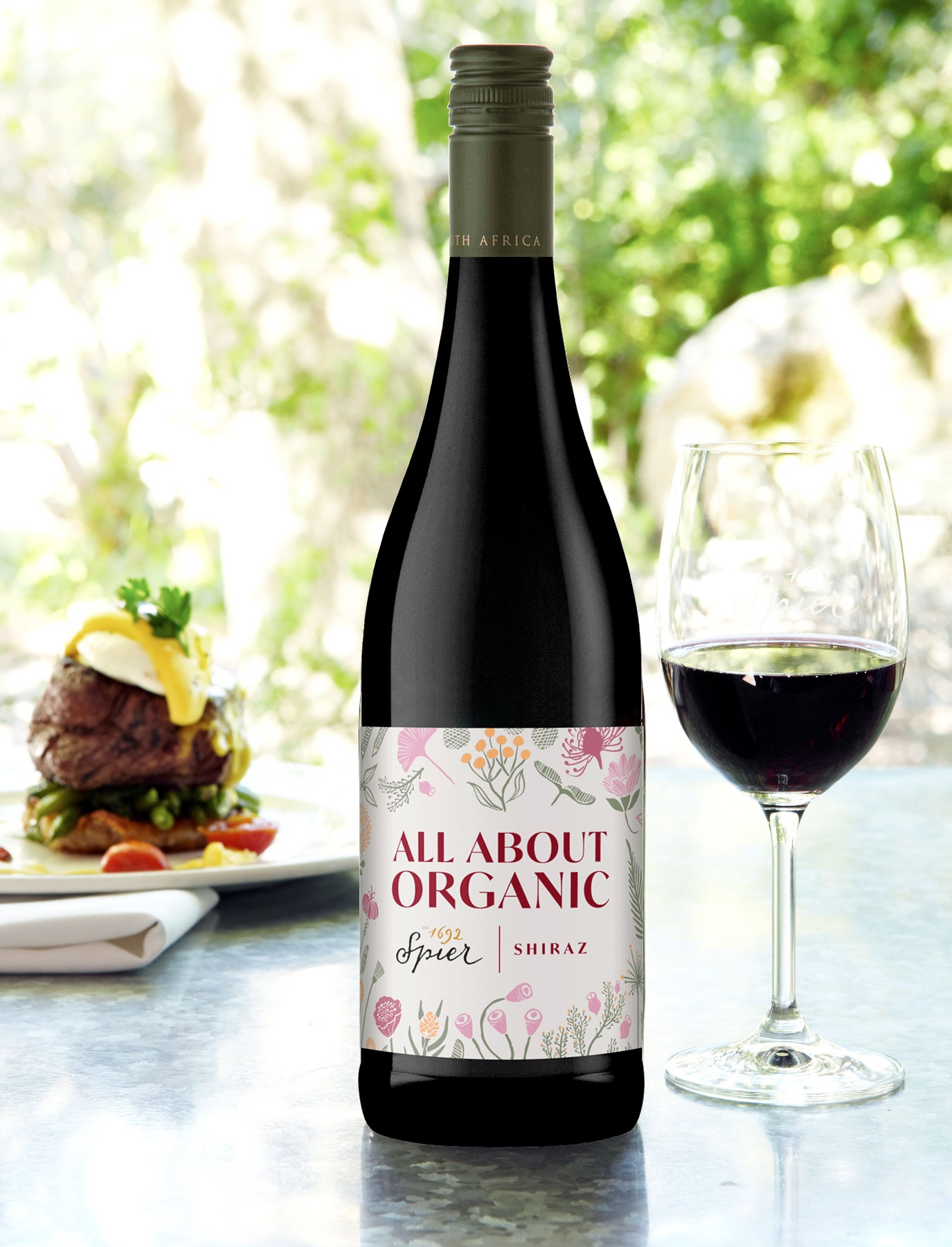 Eine Flasche Spier All About Organic Shiraz aus dunklem Glas mit grünem Verschluss und weißem Etikett mit rosa Blumenmotiven und fettem Text.