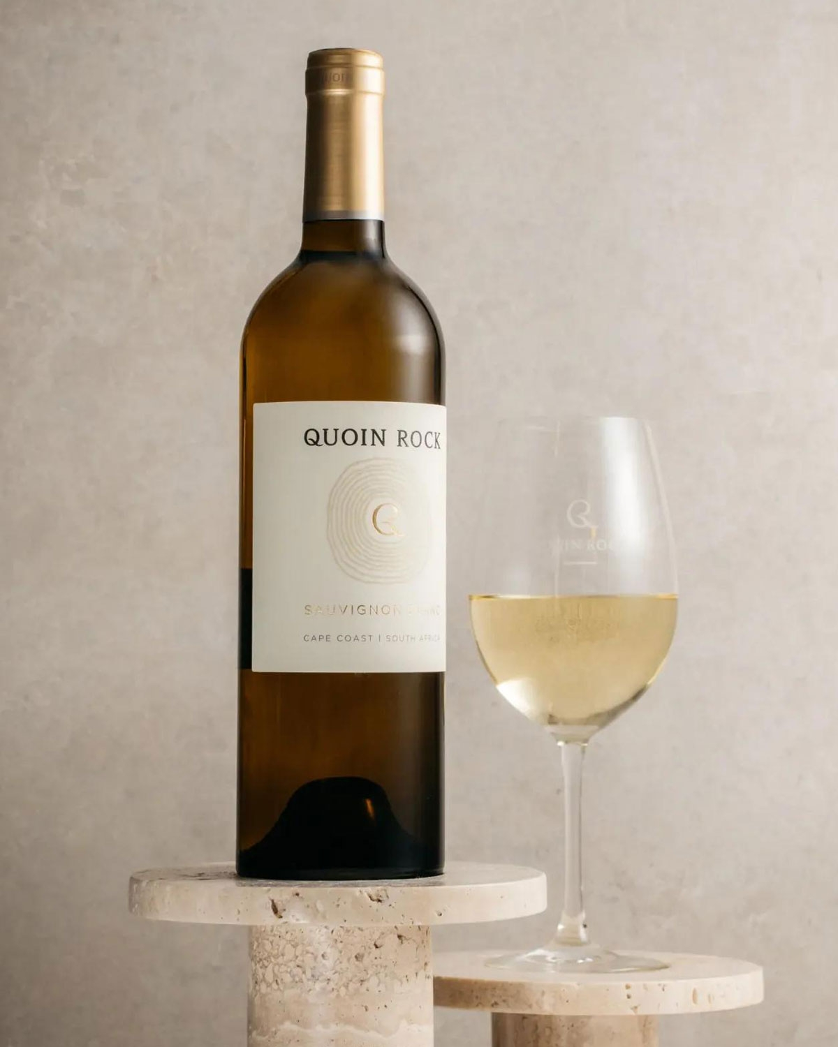 Eine Flasche Quoin Rock Sauvignon Blanc steht auf einem Marmorsockel neben einem gefüllten Weinglas, vor einem neutralen Hintergrund.