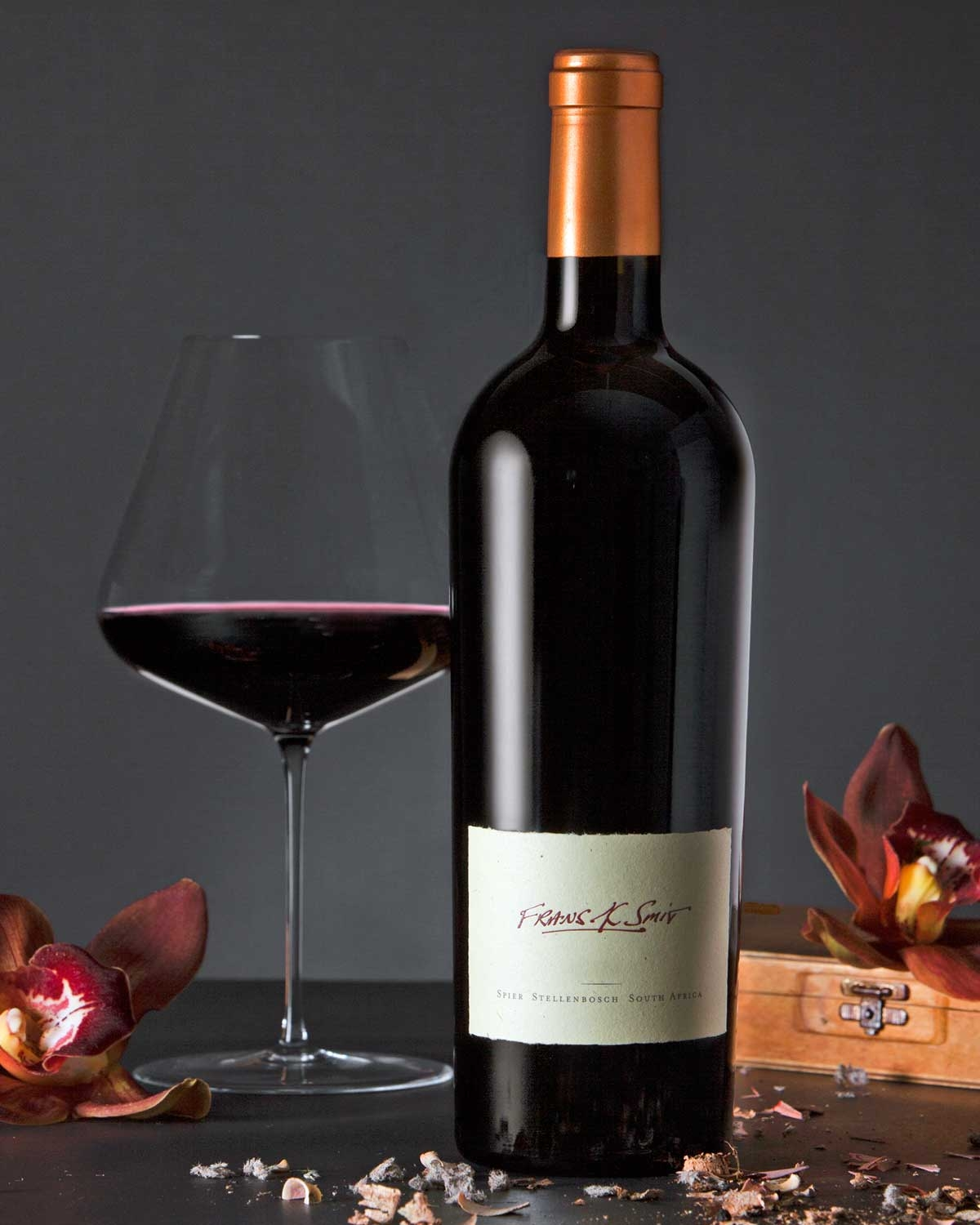 Eine dunkle Weinflasche mit einem kupferfarbenen Foliendach und einem cremefarbenen Etikett mit der Aufschrift Frans K. Smit, Hand Crafted by Spier South Africa, steht neben einer in weißes Seidenpapier eingewickelten Flasche mit einem schwarzen ovalen A