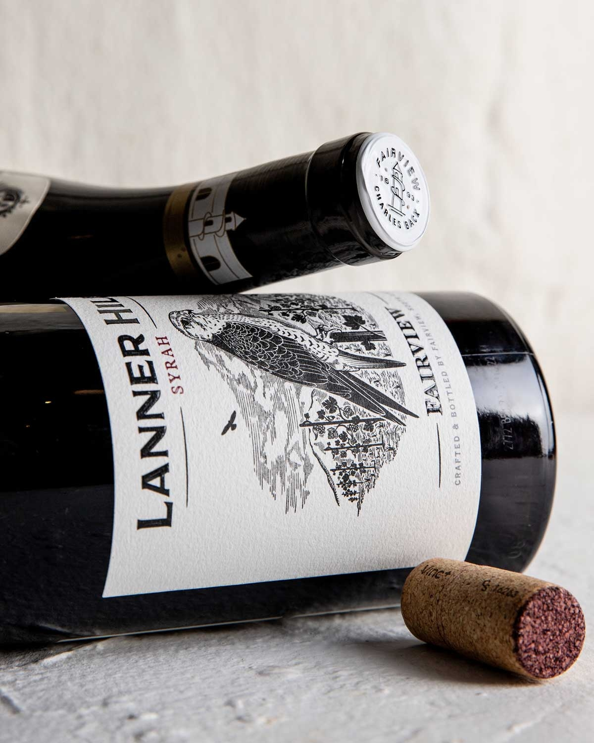 Eine Flasche des Weins Lanner Hill Syrah aus Fairview, Südafrika. Das Etikett zeigt die Illustration eines Lanner-Falken mit Bergen und Bäumen im Hintergrund und einen weißen Kapseldeckel mit einer Gebäudeillustration.