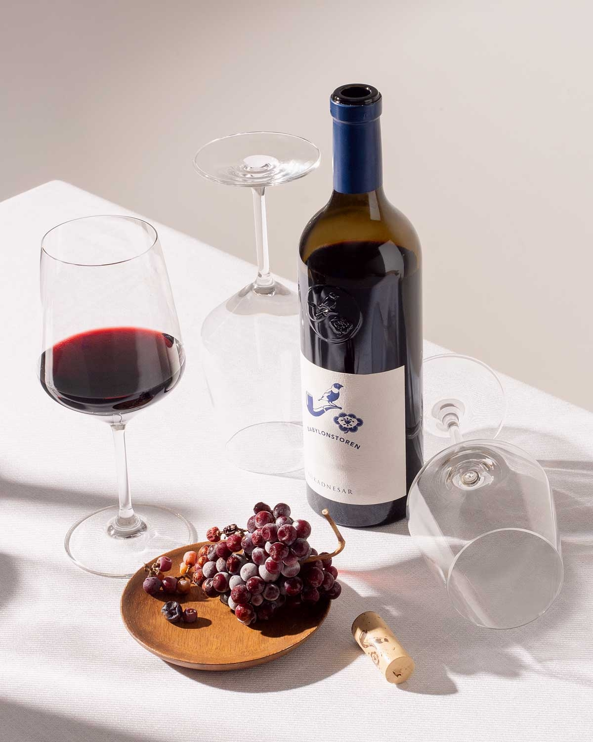Eine Flasche Rotwein, ein gefülltes Weinglas, zwei leere Weingläser, ein Korken und ein Holzteller mit Weintrauben stehen auf einem weißen Tischtuch in hellem, natürlichem Licht.