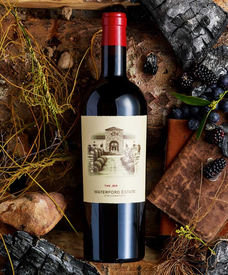 Eine Flasche Waterford Estate Wein mit rotem Foliendach liegt auf einer rustikalen Fläche, umgeben von Weintrauben, Brombeeren, verkohltem Holz und Zweigen. Das Etikett zeigt die Illustration eines steinernen Gebäudes mit Bögen.