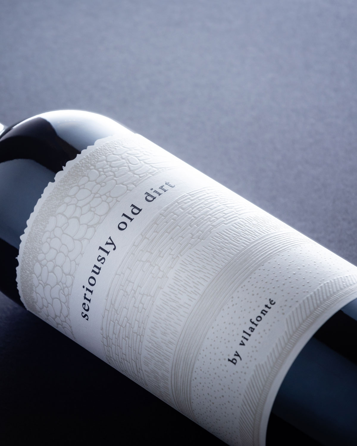 Nahaufnahme einer Weinflasche mit dem Etikett "seriously old dirt" von vilafonté mit einem geprägten weißen Etikett mit strukturierten Mustern auf dunklem Hintergrund.
